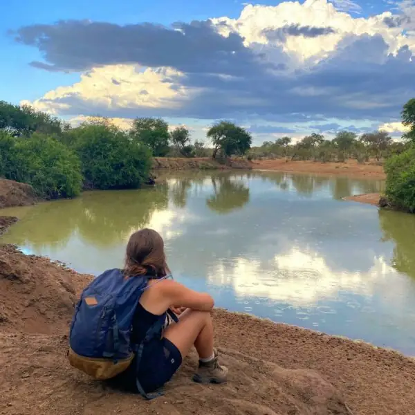 Une voyageuse assise au bord d’une rivière, contemplant les arbres qui s’y reflètent