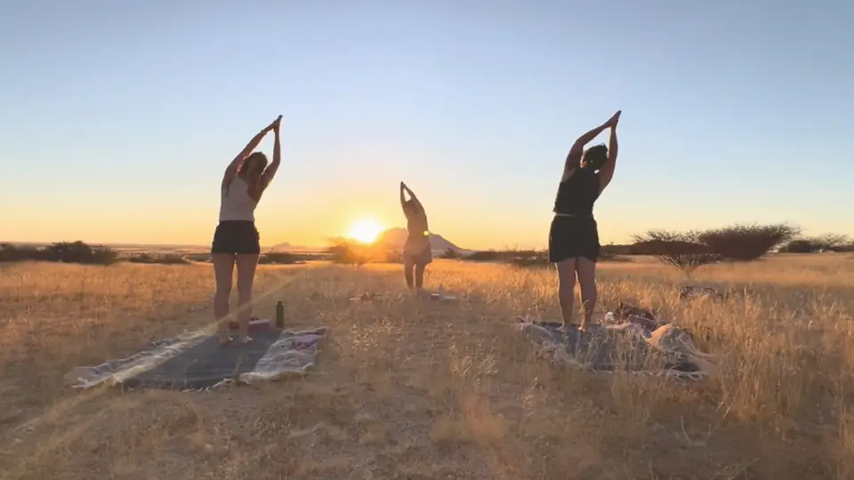 Trois femmes en posture de salutation, bras levés face au soleil couchant dans la savane africaine